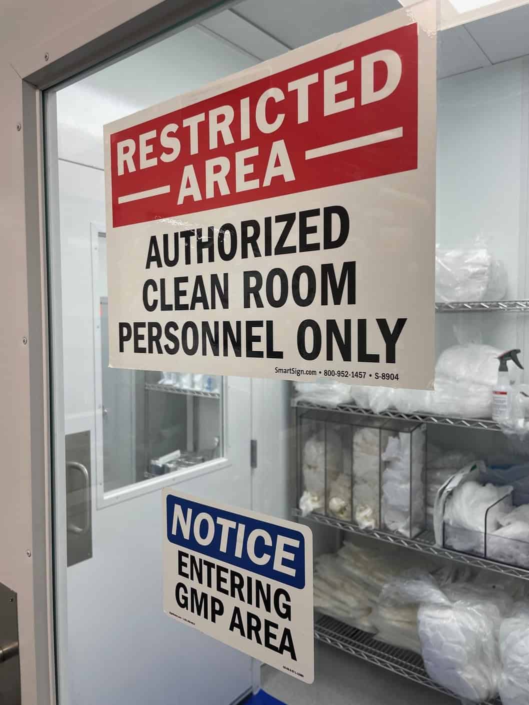 A door with signs reading "Restricted Area Authorized Clean Room Personnel Only" and "Notice Entering GMP Area," leading to a clean room with shelves holding supplies. A door with signs reading "Restricted Area Authorized Clean Room Personnel Only" and "Notice Entering GMP Area," leading to a clean room with shelves holding supplies.