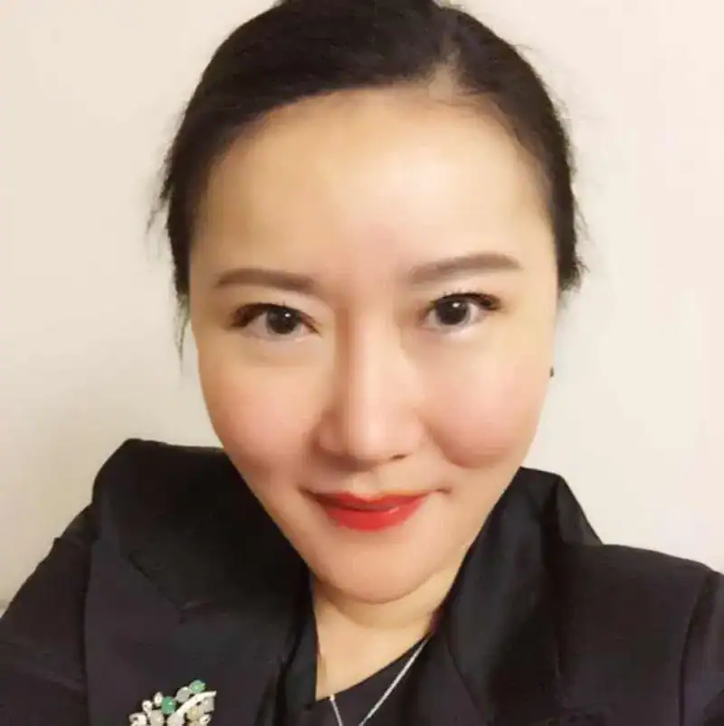 A woman with dark hair pulled back, wearing a black blazer and red lipstick, is smiling slightly in front of a plain light background.