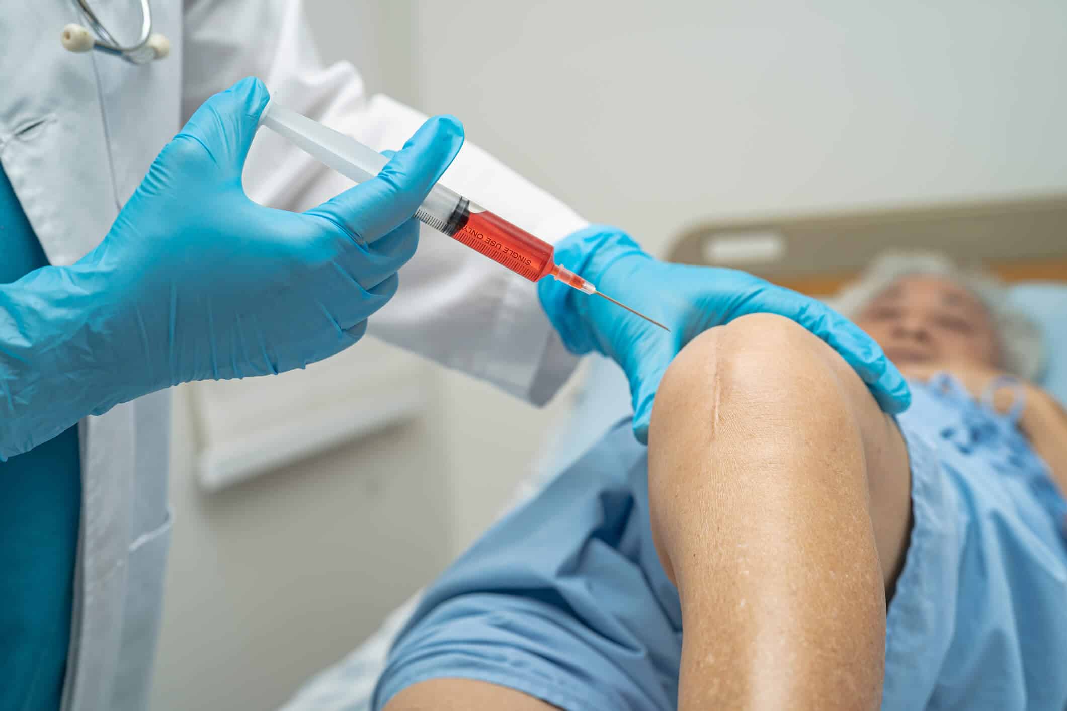 A healthcare professional prepares to inject the patient's knee with a syringe containing red fluid, possibly PRP. The patient lies on a hospital bed, wearing a blue gown.